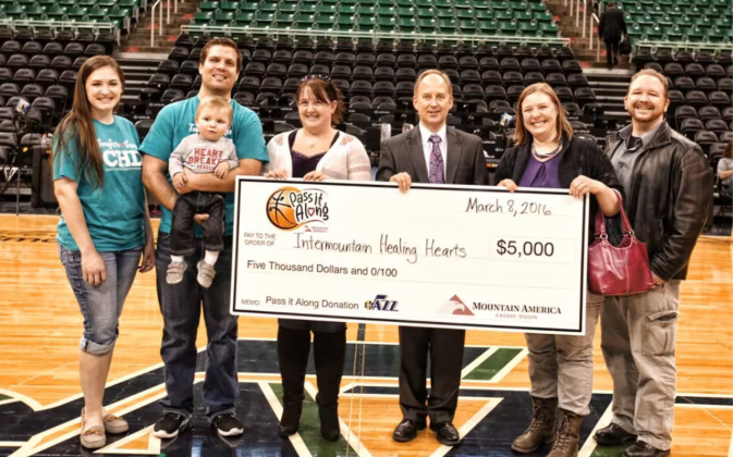 Six people stand on an arena floor holding a large ceremonial check for five thousand dollars made out to Intermountain Healing Hearts